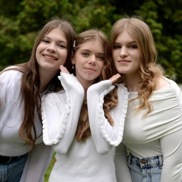 Three teenage girls in casual outfits smile while posing together outdoors in a park setting.