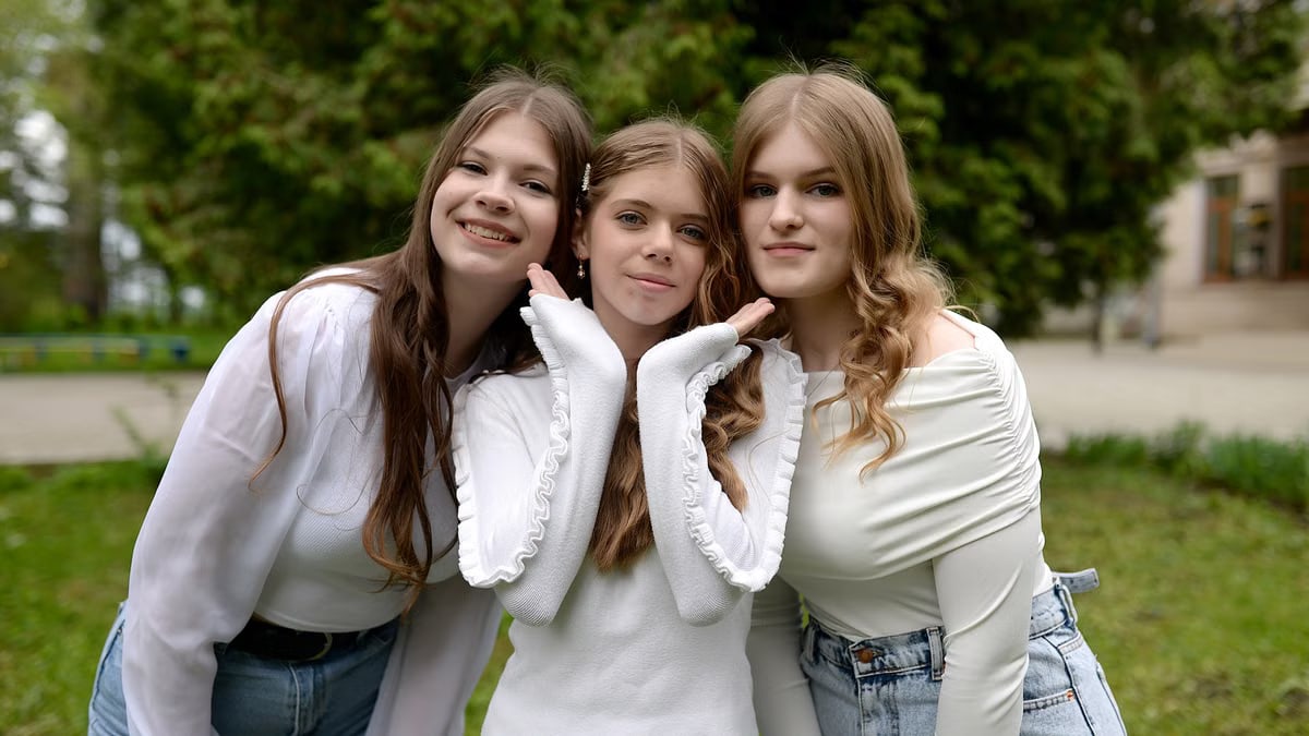 Three teenage girls in casual outfits smile while posing together outdoors in a park setting.