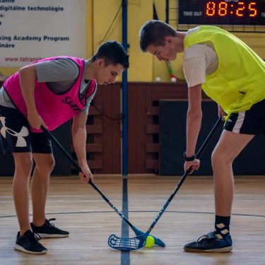 Two teenagers face off in a floorball match inside a sports gym with equipment visible in the background.